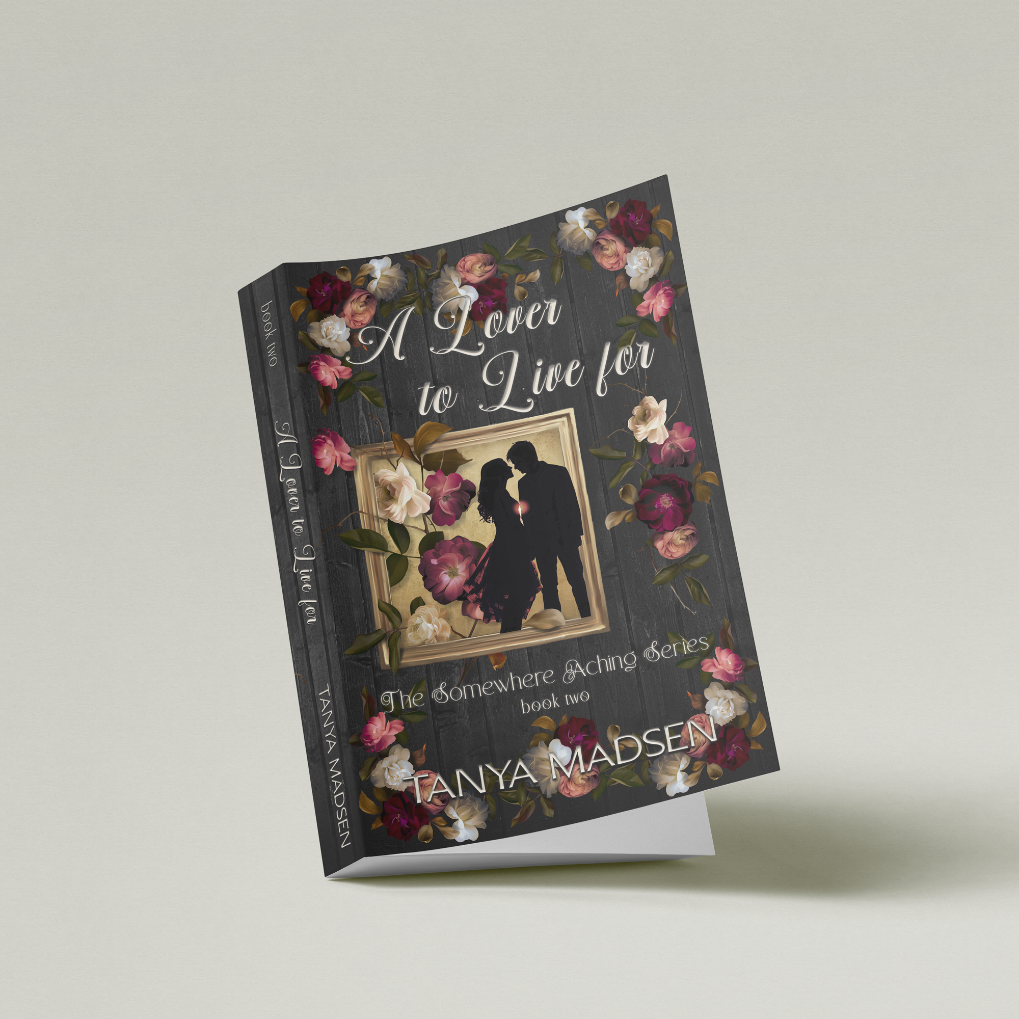 A side image of the book A Lover to Live For, surrounded by a floral design. The cover shows a silhouette of a couple almost kissing, the sun peaking between them. The book is set against a sage‑green background.