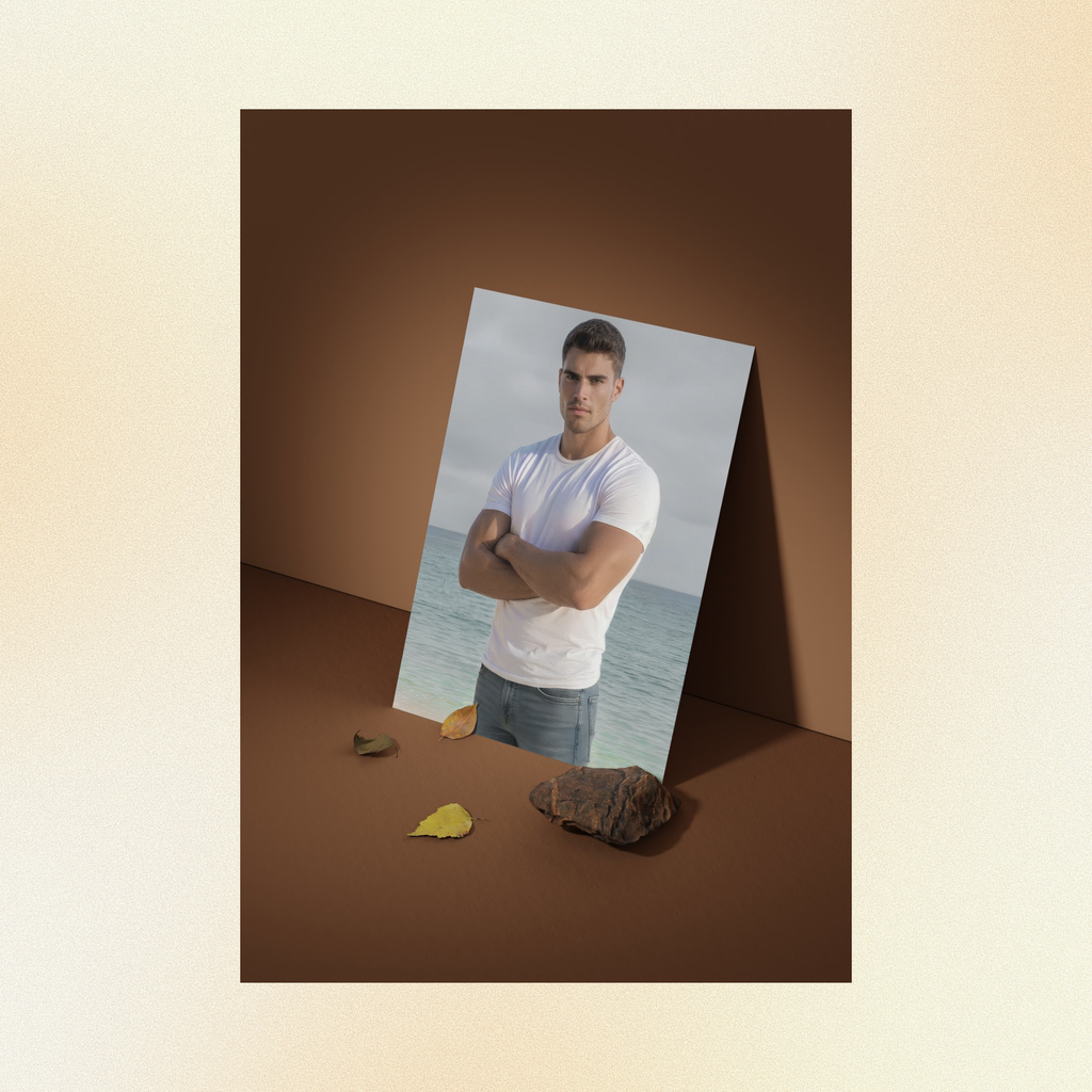 A photo of Justin displayed against a brown wall, staring intensely at the camera with his arms folded, wearing a fitted white t-shirt and faded jeans standing in front of the ocean. 