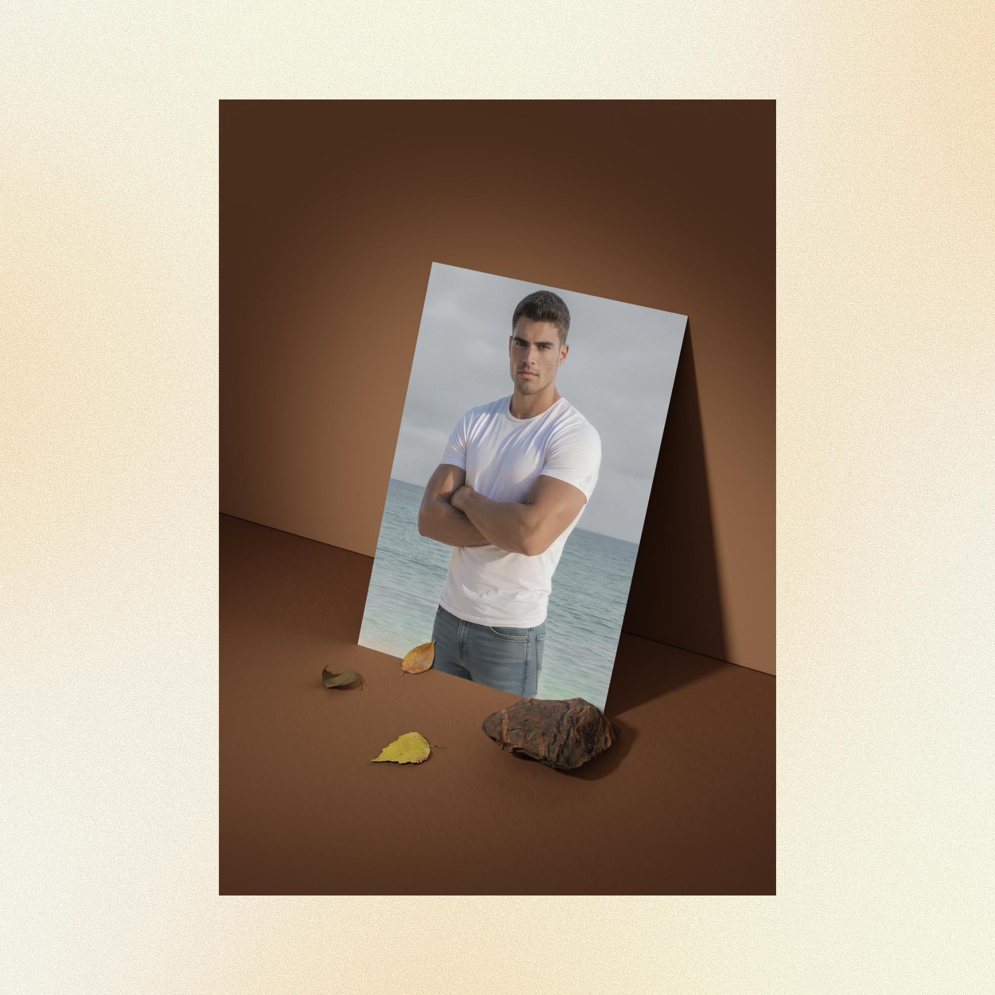 A photo of Justin displayed against a brown wall, staring intensely at the camera with his arms folded, wearing a fitted white t-shirt and faded jeans standing in front of the ocean. 