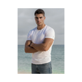 A photo of Justin staring intensely at the camera with his arms folded, wearing a fitted white t-shirt and faded jeans standing in front of the ocean. 